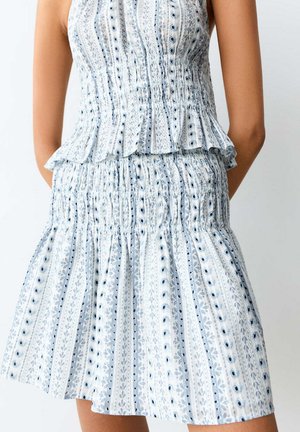 Femme portant une robe blanche sans manches smockée avec des motifs floraux bleus verticaux et à pois sur un fond uni.