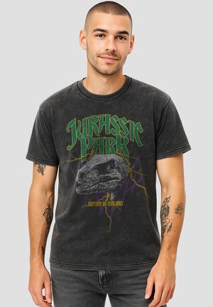 Man with short hair and tattoos wearing a faded black Jurassic Park T-shirt featuring a dinosaur head and "Raptors on Tour 1993" text.