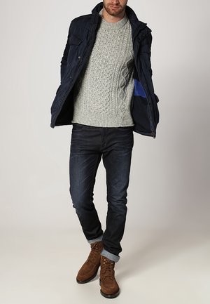 Hombre con chaqueta azul marino, suéter gris claro de punto trenzado, vaqueros oscuros con puños remangados y botas marrones de ante con cordones, de pie frente a un fondo liso.