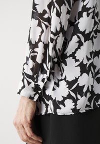 Blusa de patrón floral en blanco y negro con tela translúcida, mangas largas, puños abotonados y un corte relajado. Textura suave y ligera.