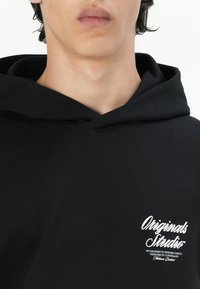 Black hooded sweatshirt with a soft texture. Features a white logo on the left chest, including text in a cursive font.
