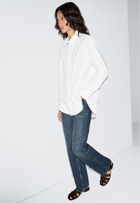 Hvid langærmet, knappet skjorte med løs pasform, kombineret med mørkeblå jeans med lige ben og brune sandaler med stropper. Enkel design, glat tekstur.