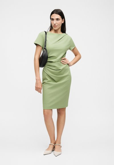 Γυναίκα με ανοιχτοπράσινο φόρεμα μέχρι το γόνατο με κοντά μανίκια, μπεζ μυτερά παπούτσια και μαύρη τσάντα στον ώμο, στέκεται με το χέρι στη μέση.