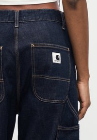 Person iført mørkeblå Carhartt jeans med synlige baklommer og kontraststing, vist fra midjen til midt på låret.