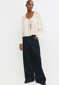 Cremfarbene Bluse aus strukturiertem Stoff, mit vorderem Bindebandverschluss und bestickten Details, kombiniert mit weit geschnittenen, dunklen Jeans und schwarzen Schuhen.