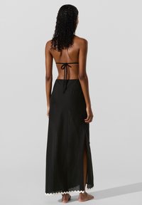 Sort maxinederdel med høj slids, lavet af letvægtsstof. Har en bølget kant og justerbare stropper med bindebånd bagpå.