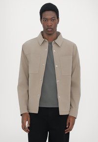 Light beige jacket with snap buttons, two chest pockets, and a collared neckline. Worn over a dark gray shirt and black pants. Smooth texture.