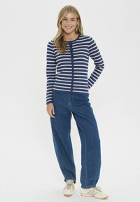 Cárdigan de botones a rayas en azul marino y crema, ajustado en el torso, combinado con unos vaqueros azules holgados y zapatillas de color claro.