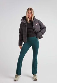 Puffet sort jakke med en hætte, en lyserød detalje på brystet, parret med teal leggings og beige sportssko. Tekstureret og tætsiddende design.