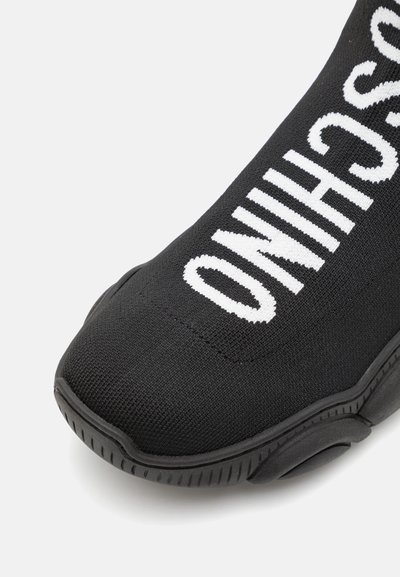 Black athletic shoe with a knitted upper featuring large white lettering. Rubber sole with textured detailing and rounded toe.