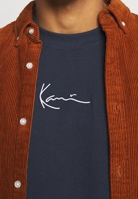 Camisa de pana marrón con botones blancos, superpuesta sobre una camiseta azul con el texto bordado en blanco "Kami" en el pecho.