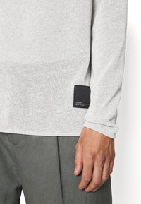 Haut à manches longues gris clair en tissu maille texturé, avec une étiquette noire en accent à l'ourlet. Main visible tenant le tissu.