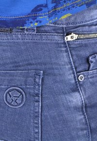 Blå denimjeans med dragkedja, med en synlig dragkedja, fickstickningar och en cirkulär logotyp broderad på framsidan av fickan.
