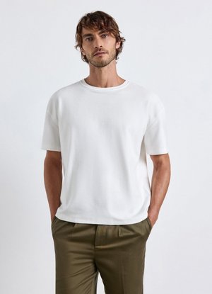 Man with curly hair wearing a plain white short-sleeve shirt and olive green pants standing against a white background.