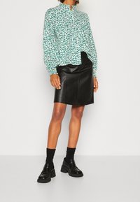 Grün-weiße gemusterte Bluse, lange Ärmel mit elastischen Bündchen, kombiniert mit einem schwarzen Leder-Bleistiftrock und klobigen schwarzen Schuhen.