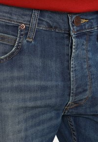 Vaqueros de denim de color azul oscuro, con costuras visibles, remaches y un pequeño logotipo de la marca en la cintura. La textura parece suave.