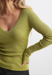 Una donna che indossa un pullover attillato a coste verde lime con maniche lunghe e scollatura a V, con una mano appoggiata sui fianchi su uno sfondo neutro.