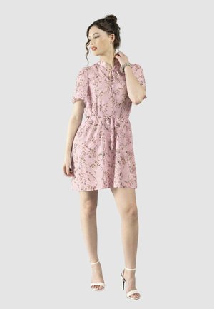 Donna che indossa un vestito corto a maniche corte con stampa floreale rosa e cintura in vita, sandali bianchi con tacco alto e un braccialetto, posando con la mano vicino al collo.