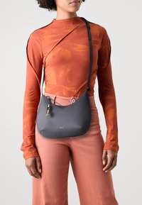 Borsa a tracolla in pelle grigia con una forma strutturata, dettagli in metallo dorato e tracolla rimovibile, indossata sopra una maglietta arancione a maniche lunghe e pantaloni rosa.