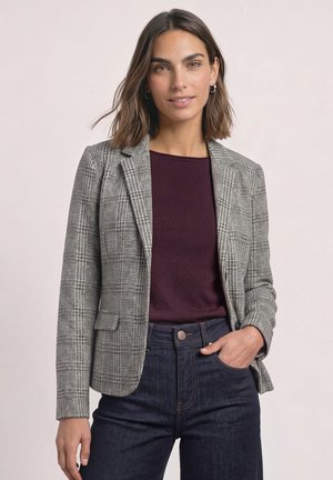 Femme aux cheveux bruns mi-longs porte un blazer à carreaux gris, un haut bordeaux et un jean bleu foncé, posant avec une main dans sa poche.