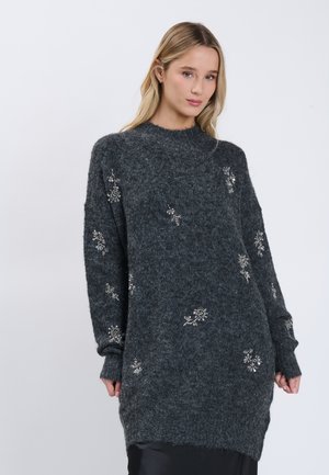 Jonge vrouw draagt een donkergrijze zachte sweaterjurk met zilveren bloemrijke borduurwerk en poseert tegen een effen lichte achtergrond.
