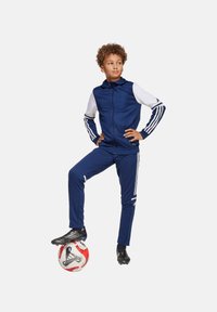 Blauwe sportieve trainingspak met witte mouwen, voorzien van drie strepen langs de armen en broek. Kind staat op een rood-witte voetbal.