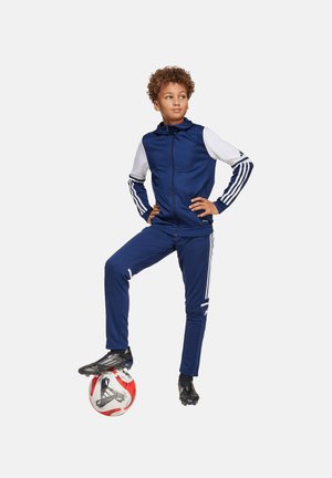 Blauer Sportanzug mit weißen Ärmel, der drei Streifen entlang der Arme und Hosen hat. Ein Kind steht auf einem rot-weißen Fußball.