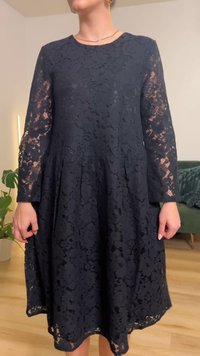 Vestido preto de renda com mangas compridas, corte evasê, decote redondo, padrão floral e tecido texturizado. O comprimento do vestido fica ligeiramente abaixo do joelho.