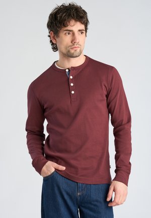 Man with dark curly hair wearing a long-sleeve maroon henley shirt and blue jeans, standing with one hand in pocket, looking to the side.