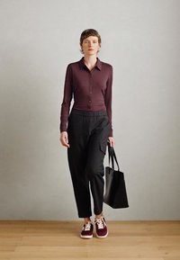 Chemise à manches longues bordeaux, pantalons noirs texturés, baskets bordeaux et sac fourre-tout noir, debout sur un sol en bois devant un fond gris.