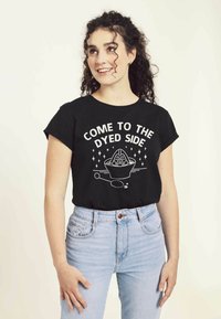 Camiseta de algodón negra con mangas cortas que presenta texto en blanco y un diseño gráfico. Se lleva con jeans de cintura alta de color azul claro.