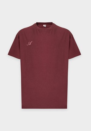 Άνδρας που φοράει μπορντό T-shirt Arkk Copenhagen με ροζ γράμματα και μικρό κείμενο στην πλάτη, στέκεται μπροστά σε απλό λευκό φόντο.