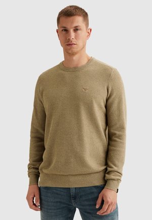 Jonge man die een beige fijngebreide trui met lange mouwen en een blauwe spijkerbroek draagt, staand tegen een effen lichte achtergrond.