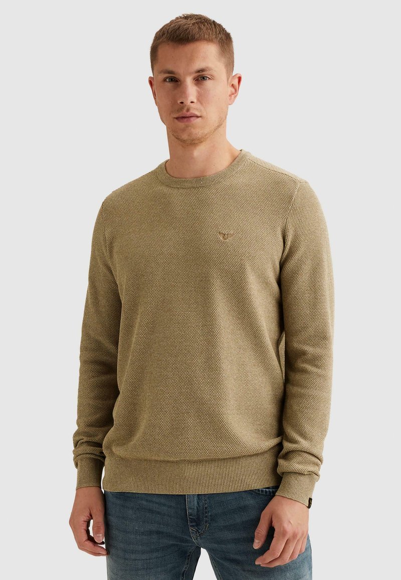 Jeune homme portant un pull à manches longues beige texturé et un jean bleu, debout devant un fond clair uni.