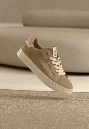 Beige low-top sneaker with cream laces and sole, decorative scalloped edge, and "1978" label on the tongue, floating above textured carpet.