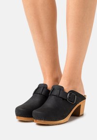 Sabots en cuir noir avec une finition texturée, bout rond, talon en bois et détail de boucle sur le côté pour un ajustement réglable.