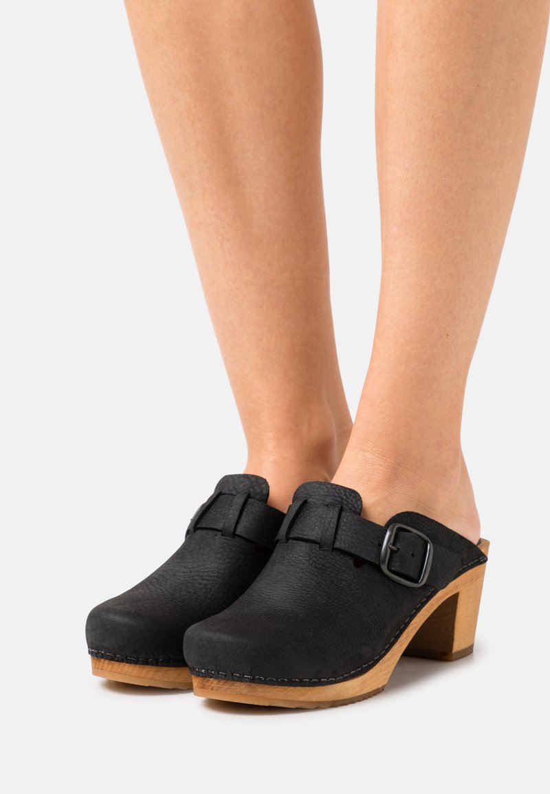 Sabots en cuir noir avec une finition texturée, bout rond, talon en bois et détail de boucle sur le côté pour un ajustement réglable.