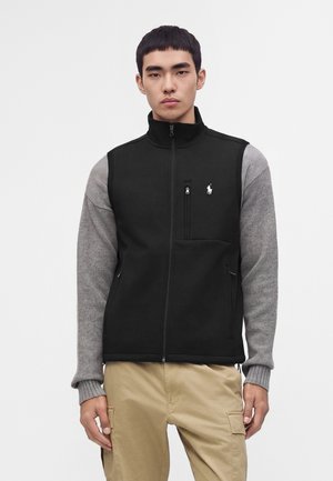 Jeune homme portant un gilet noir zippé par-dessus un pull gris à manches longues et un pantalon beige, debout devant un fond blanc uni.
