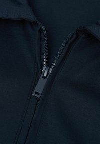 Czarna tkanina w zbliżeniu, ukazująca gładki zamek błyskawiczny z plastikowym uchwytem, otoczony przeszytymi krawędziami i lekko teksturowaną powierzchnią.