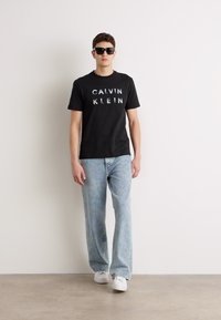 Tricou negru din bumbac cu logo-ul "CALVIN KLEIN", blugi din denim albastru deschis, adidași albi și ochelari de soare negri, pe un fundal neutru.