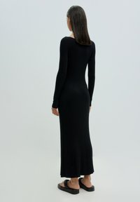 Schwarzes, geripptes Strickkleid mit langen Ärmeln und figurbetontem Schnitt, das knielang ist und einen runden Rückenausschnitt hat, getragen mit Pantoletten.