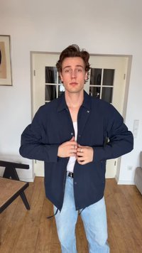 Jeune homme debout à l'intérieur, boutonnant une veste bleu marine sur une chemise blanche, portant un jean bleu clair, avec un sol en bois et une porte vitrée derrière.