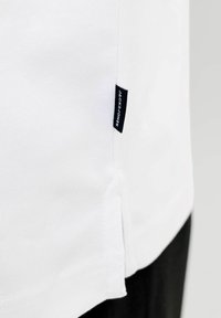 Tissu blanc avec une petite étiquette noire cousue dans la couture, affichant le nom de la marque "Jack & Jones" en texte blanc.