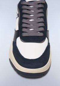 Sneaker con una toma sintetica color crema, accenti in suede blu navy, lacci grigi e una suola in gomma beige. Presenta una punta arrotondata e una linguetta imbottita.
