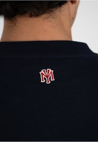 Vue de dos d'une personne portant un pull à col côtelé bleu marine avec un petit logo « NY » rouge et blanc brodé, centré sous le col.