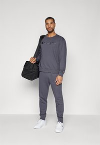 Grauer Pullover mit "Champion"-Logo, passende graue Jogginghose und weiße Sneakers. Schwarze Sporttasche mit Netzdetaillierung. Einfacher und stimmiger Look.