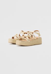 Sandales en plateforme métalliques dorées avec des brides croisées et une semelle en wedge en jute. Présente une surface légèrement texturée et un accent de boucle.