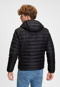 Svart puffjacka med huva, med kanalstickning och en slät, glänsande yta. Bärs tillsammans med blå denimjeans.