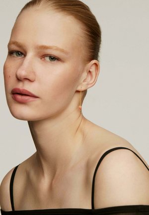 Pendientes de cadena dorada con acentos florales rojos, colgando del lóbulo de la oreja. La piel suave y el maquillaje mínimo complementan una blusa negra de tirantes.