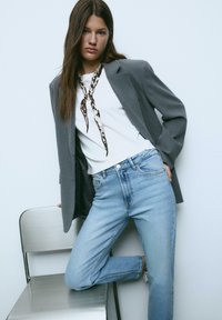 Grå skräddarsydd blazer över en vit skjorta, i kombination med ljusblå jeans. Håret är långt och rakt; en mönstrad scarf är knuten runt halsen.